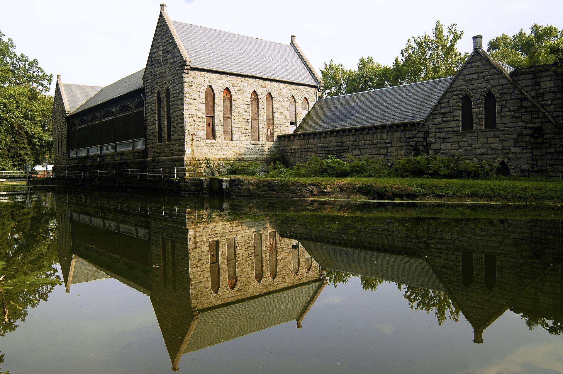 Stone building with arched windows reflected in calm water on a bright day.