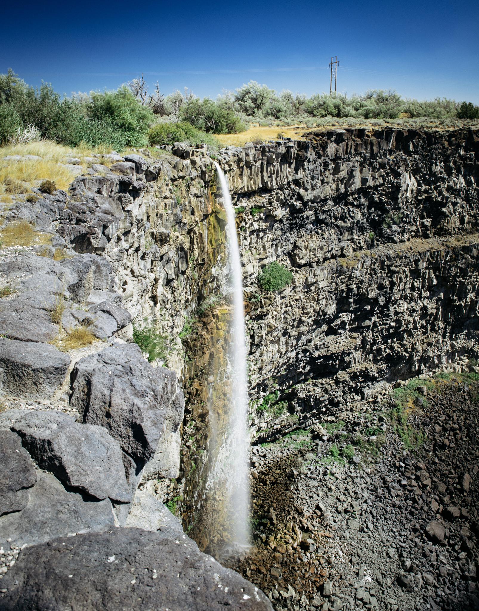 Tall waterfall plunges over a rocky cliff under a bright blue sky.