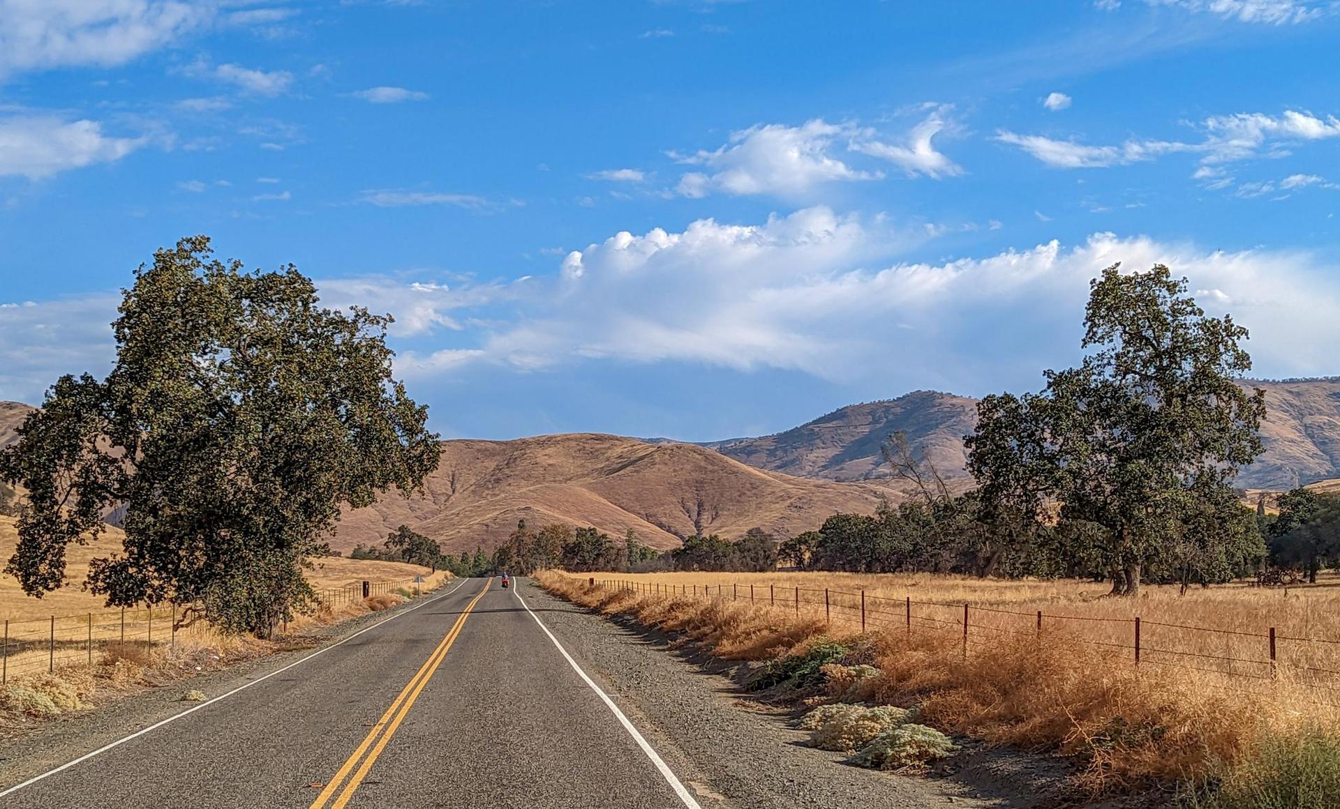 Paved road through dry fields, flanked by trees, leading to distant mountains.