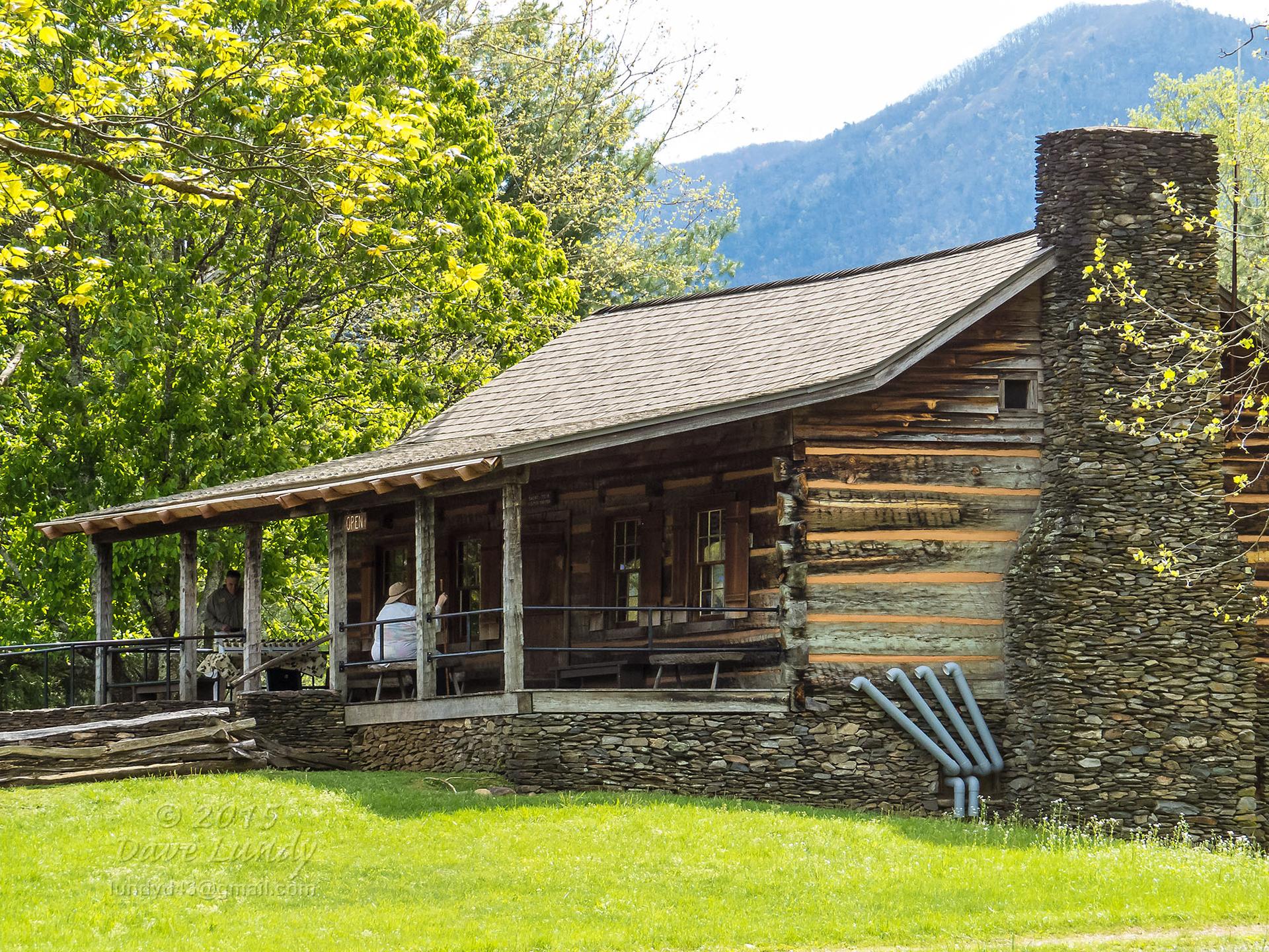Historic log cabin with a stone chimney and porch, people sitting, bright day.