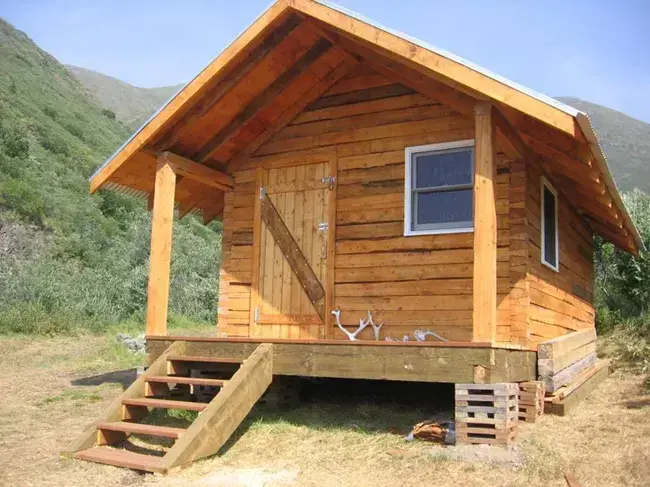 Small wooden cabin with a porch and steps, set against a green mountain.