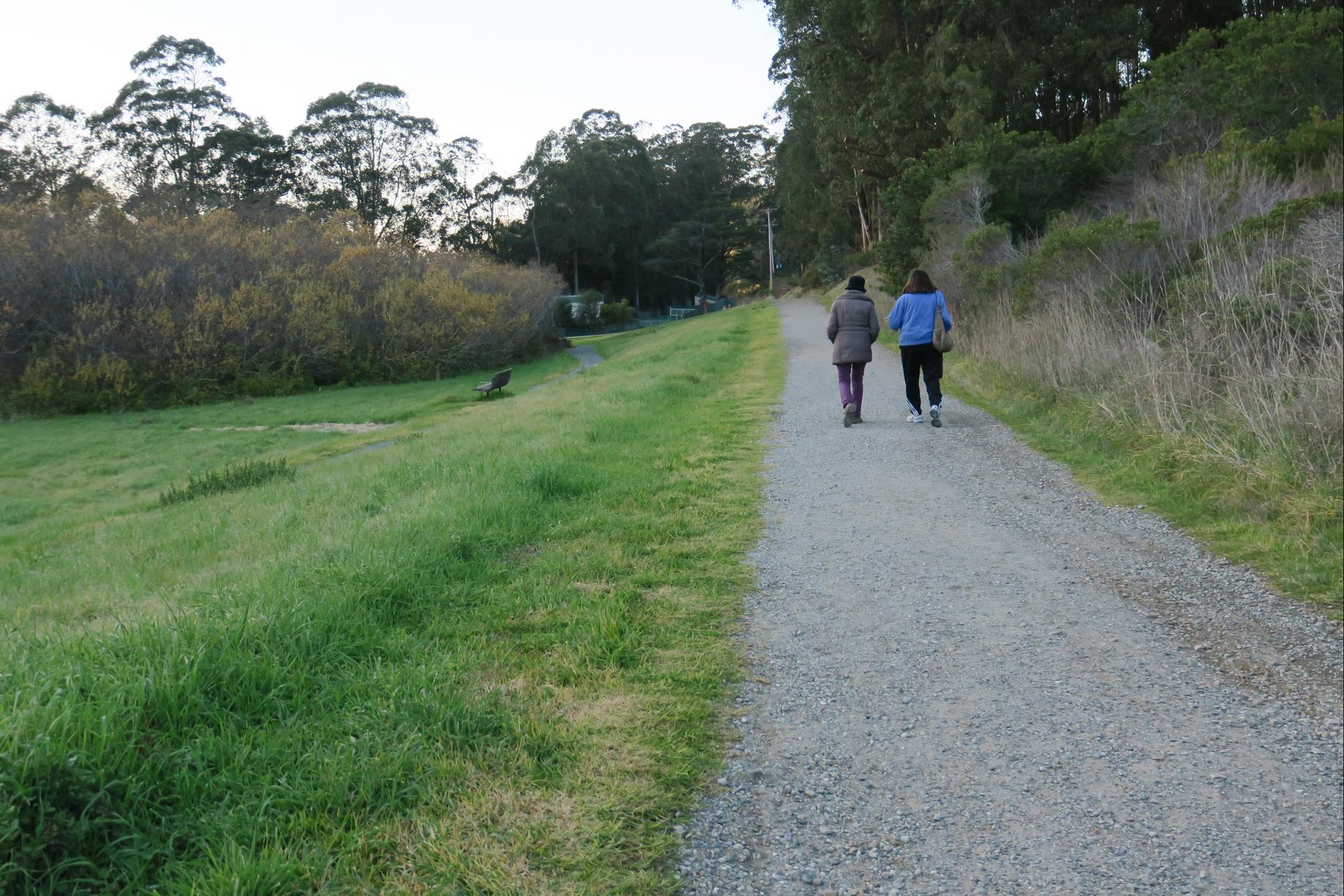 Two people walk on a gravel path beside a grassy slope and trees.