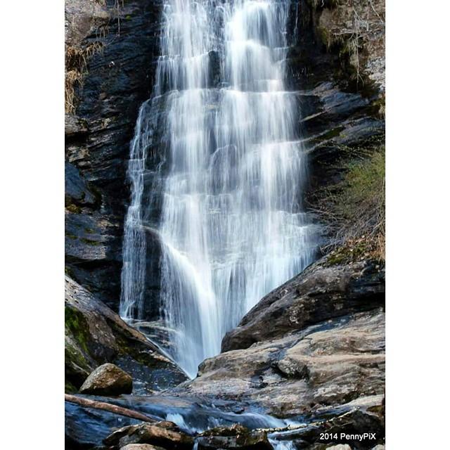 A multi-tiered waterfall cascades down dark, rocky cliffs in a natural setting.