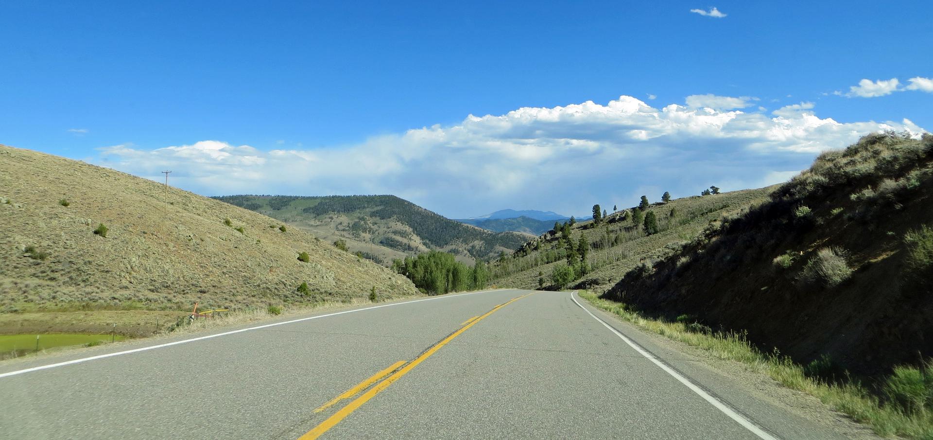 A road winds through green hills and trees under a partly cloudy sky.
