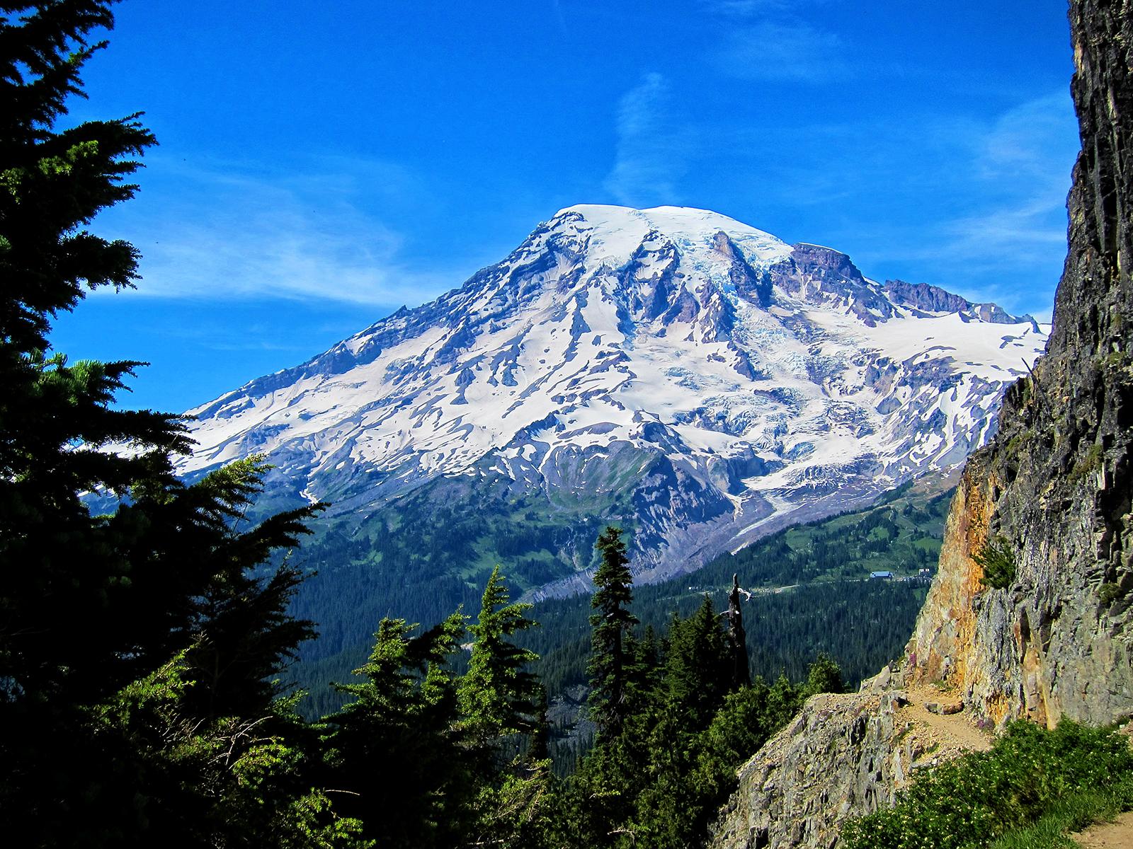 A majestic snow-capped mountain under a bright blue sky, surrounded by forests.