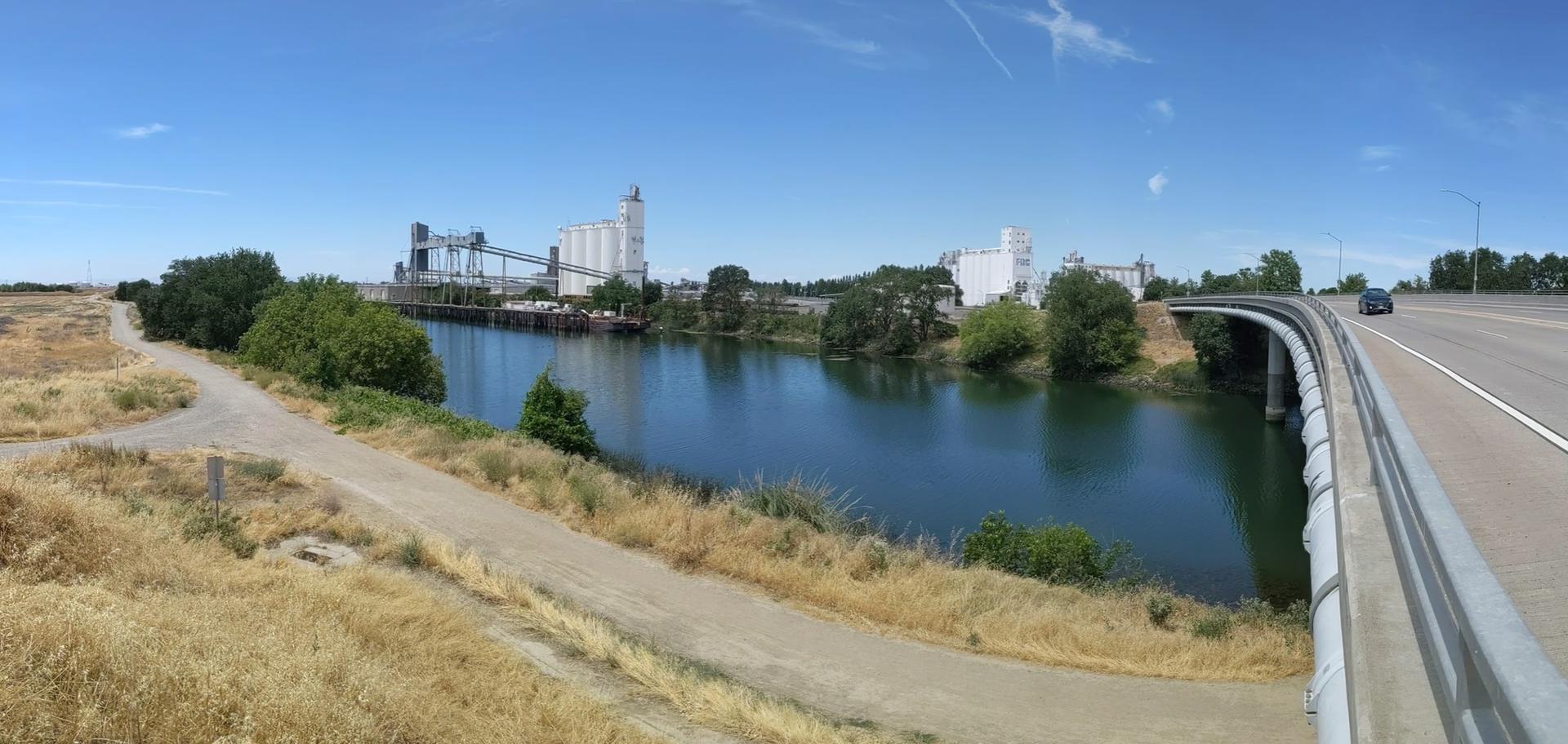 River, industrial buildings, and highway bridge under a bright blue sky with a car.