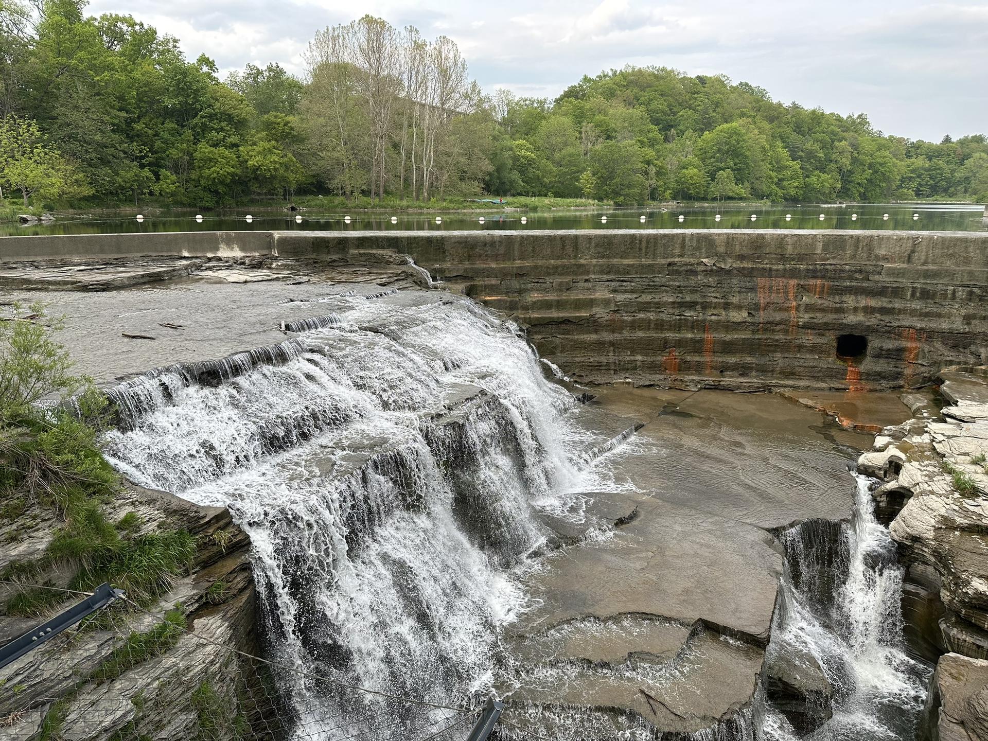 Cascading waterfall over concrete structures and rocks, surrounded by green trees under an overcast sky.
