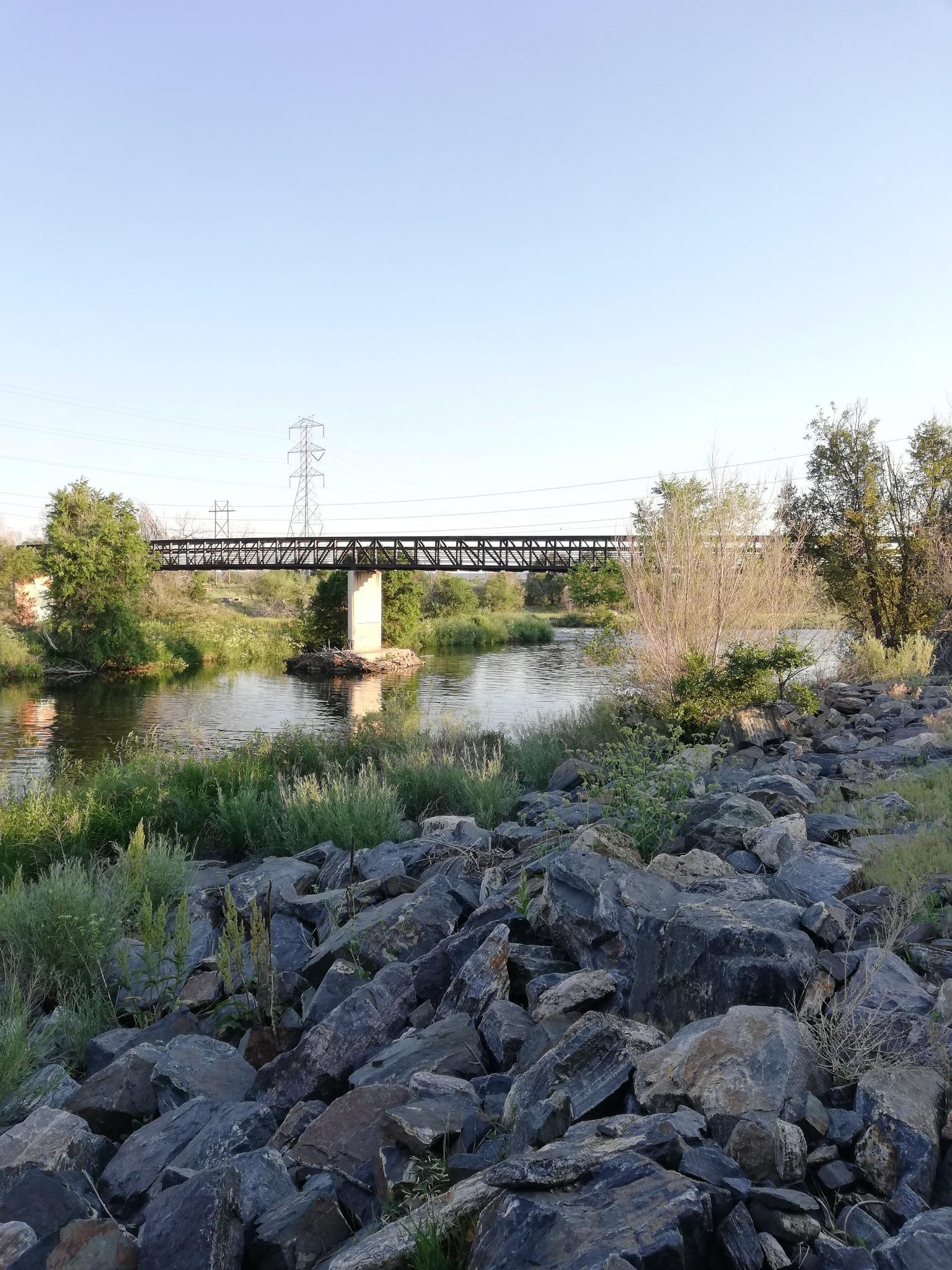A metal bridge spans a calm river with rocky banks and green trees.