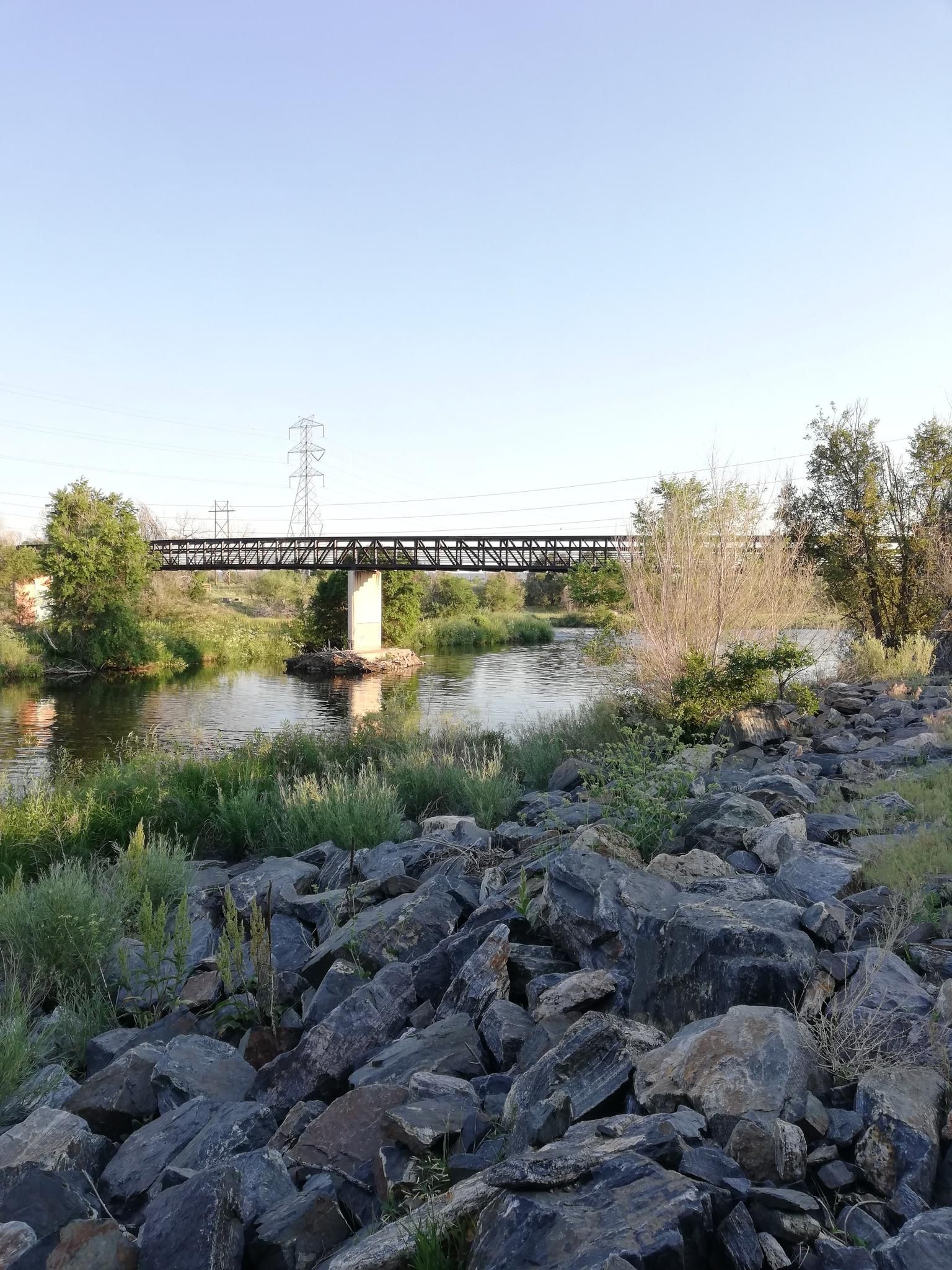 A metal bridge spans a calm river with rocky banks and green trees. - South Platte River – Lowell Ponds Wildlife Area loop from Theatre District / Convention Center