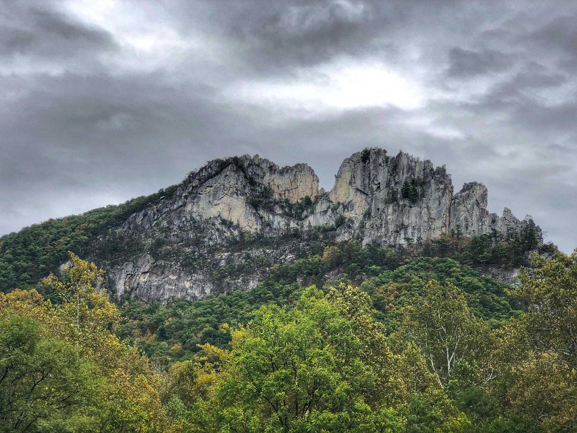 Dramatic rocky mountain ridge rises above green forest under a cloudy sky.