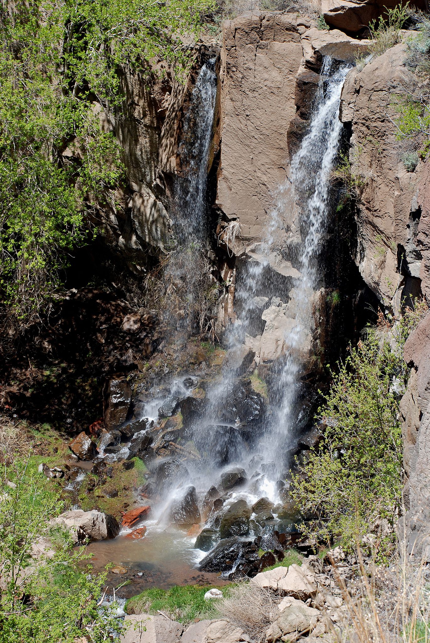 Two waterfalls cascading over dark rocks into a rocky pool. - Falls Trail