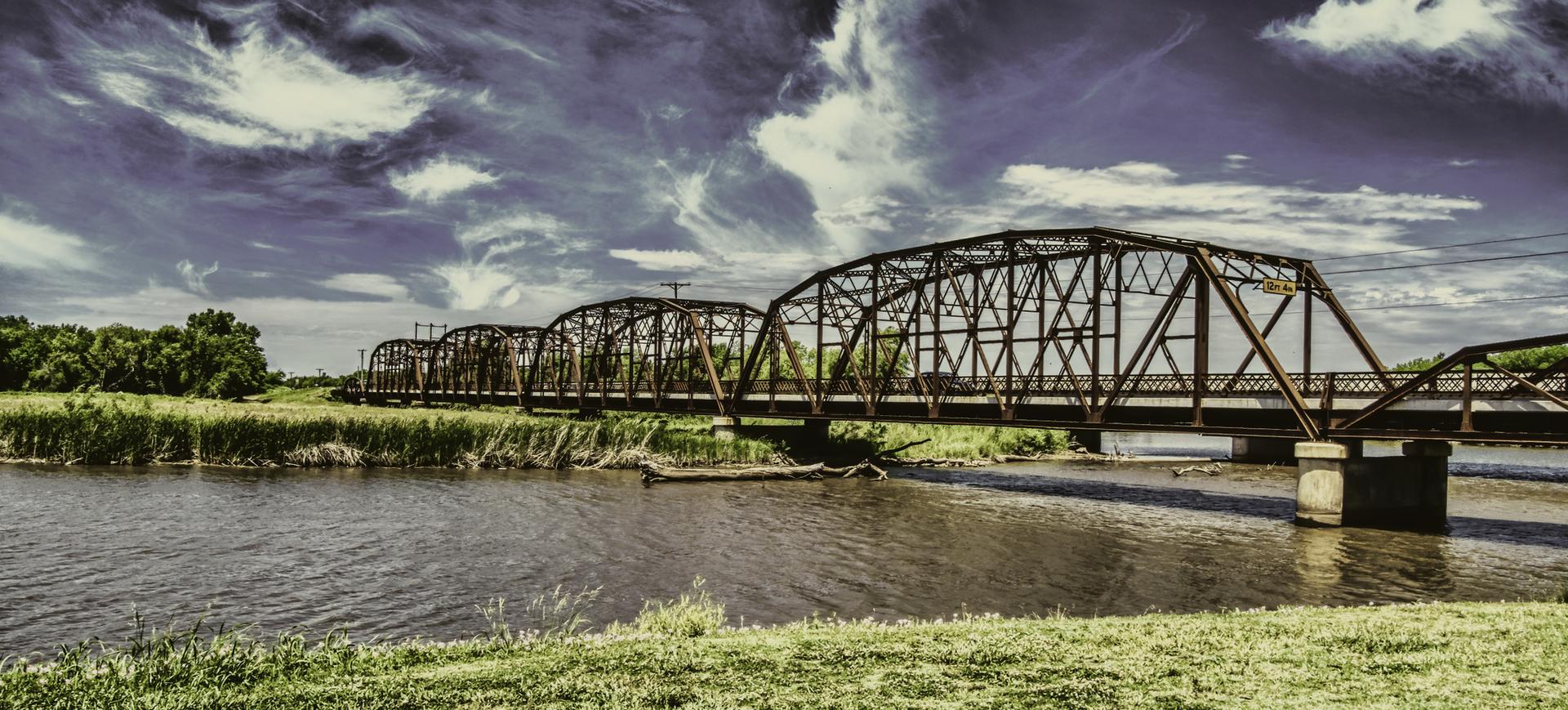 Multiple metal truss bridges span a river with green banks under a dramatic sky.