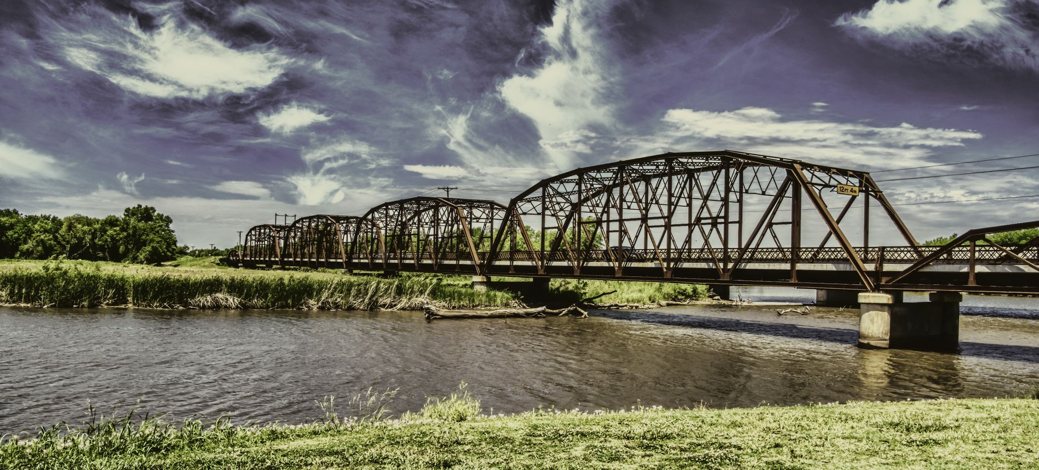 Multiple metal truss bridges span a river with green banks under a dramatic sky. - Lake Overholser Bridge – Lake Overholser Dam loop from First Indian Pentecostal Church