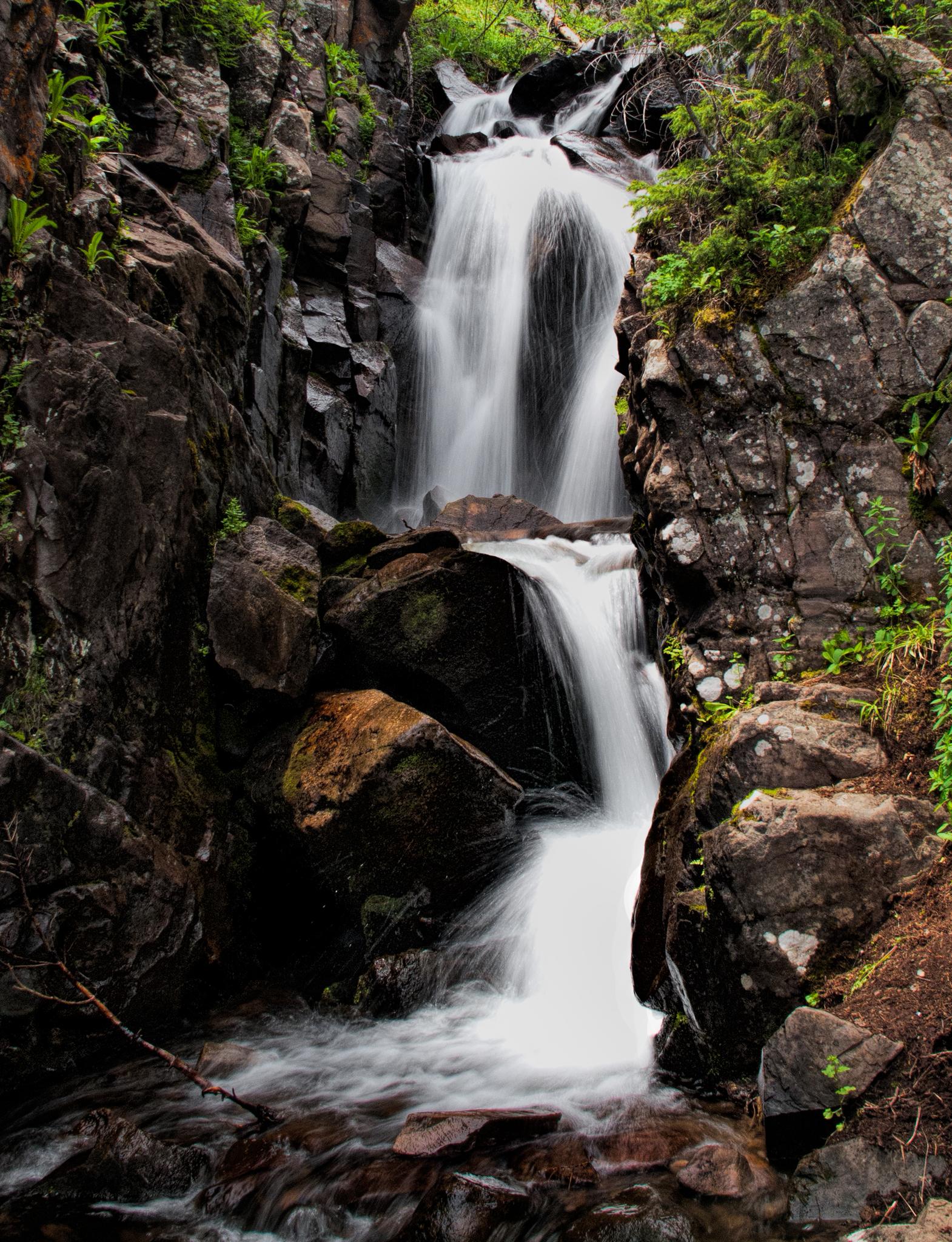 A multi-tiered waterfall cascades down dark, mossy rocks in a lush gorge.