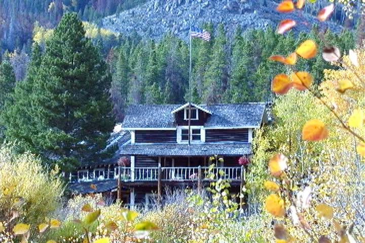 Rustic lodge with a flag, surrounded by autumn trees and a forested mountain.