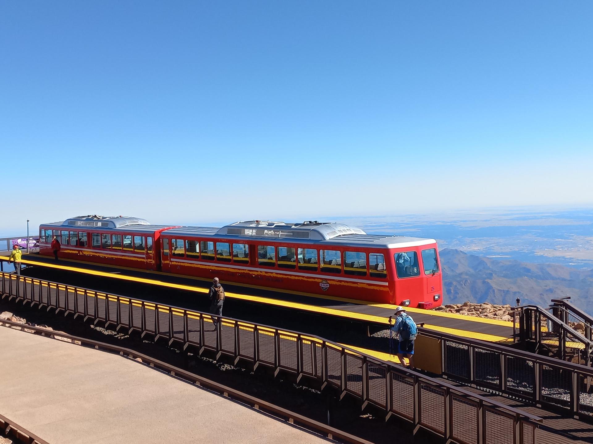 Red and silver train at a mountain station with people and clear sky.