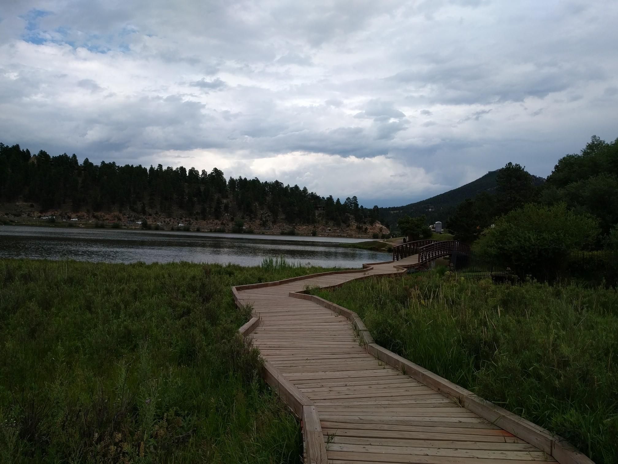 A wooden boardwalk winds through marshland beside a calm lake and forest. - Evergreen Lake – Alderfer Ranch loop from Evergreen