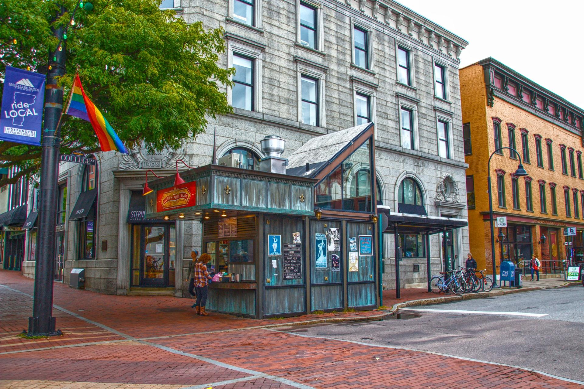An urban street corner with historic buildings, a kiosk, and people on an overcast day.
