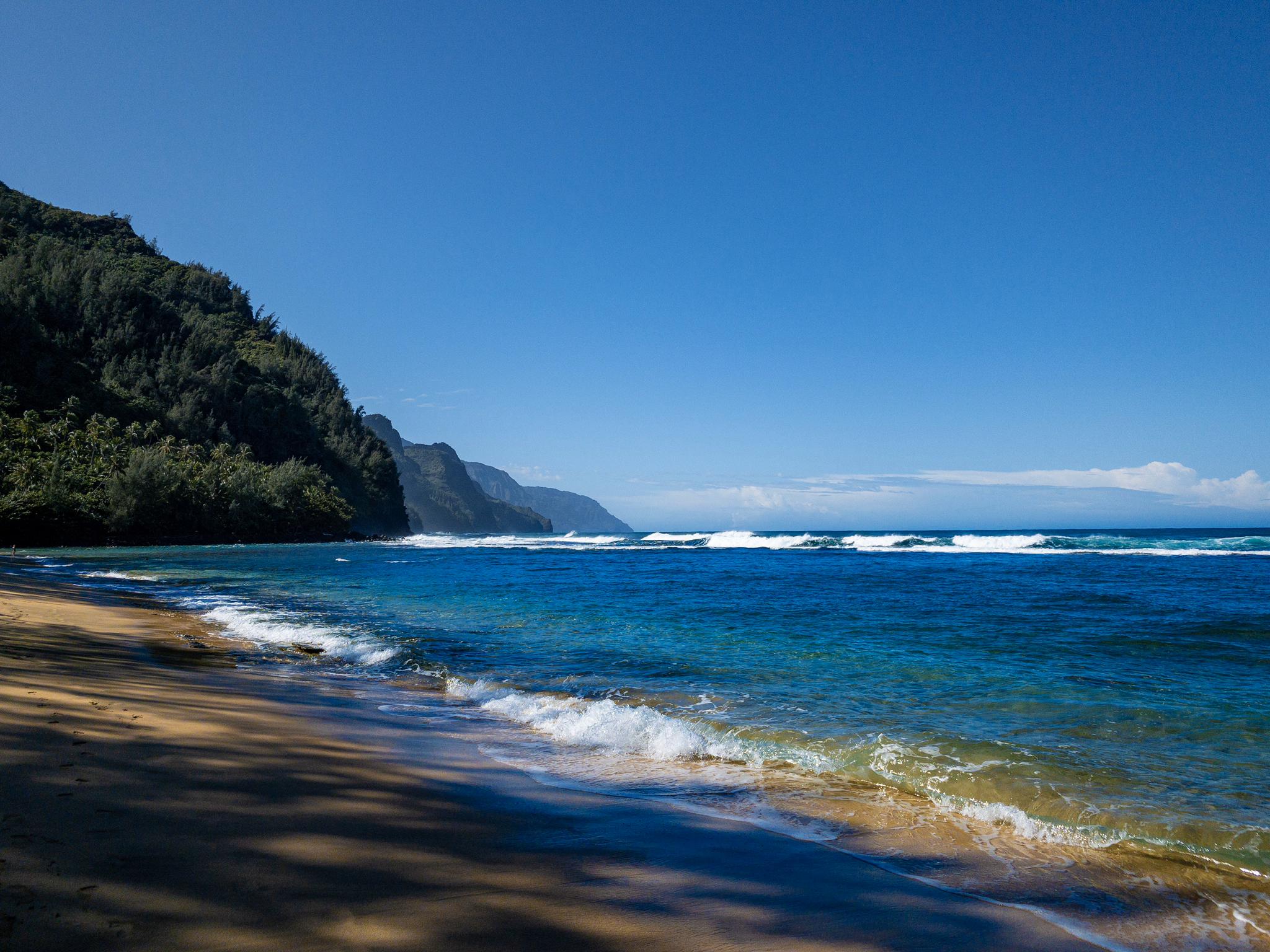 Sunny beach with gentle waves, backed by a lush green mountain.