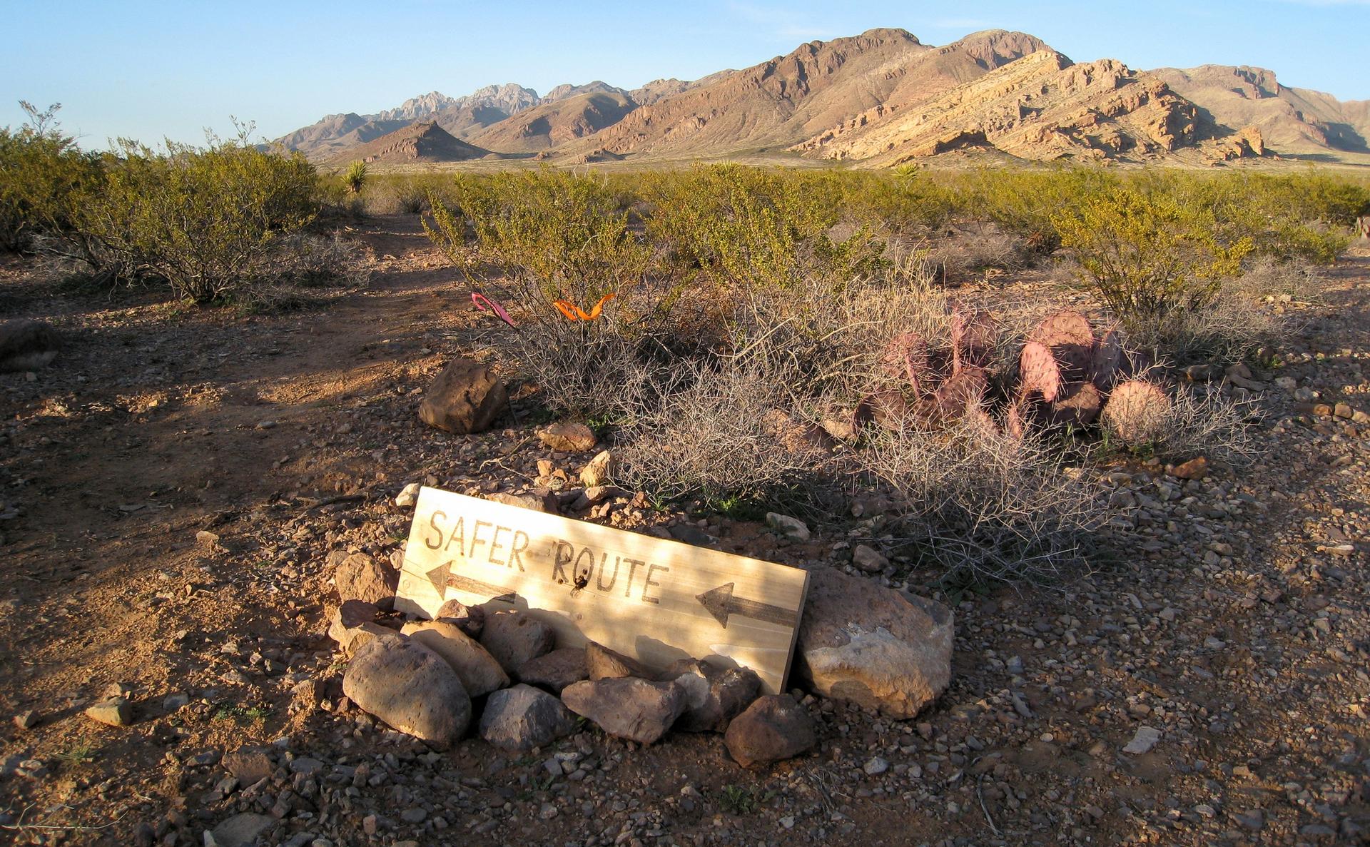 A 'SAFER ROUTE' sign on the ground in a desert landscape at sunset.
