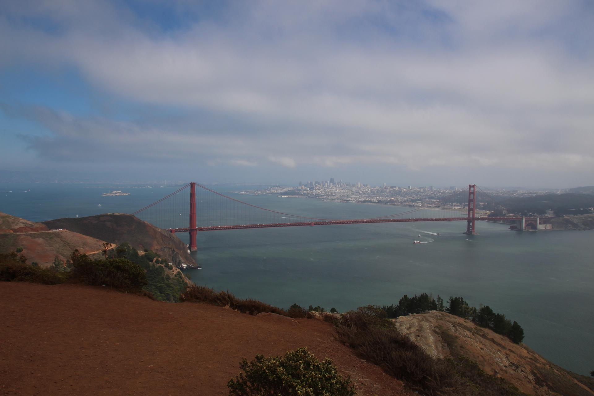 Red suspension bridge spans water, distant city skyline under a cloudy sky.