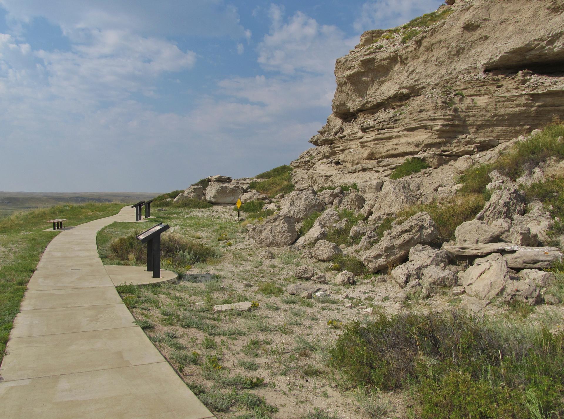 Paved pathway alongside a rocky cliff under a bright, cloudy sky.
