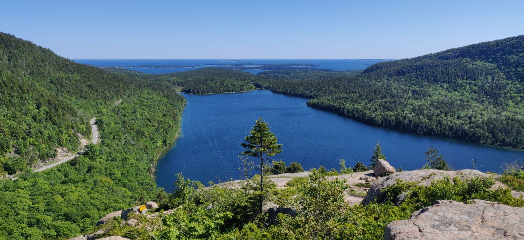 Panoramic view of a blue lake nestled between green forested mountains under a clear sky. - Jordan Pond Six-Peak Loop