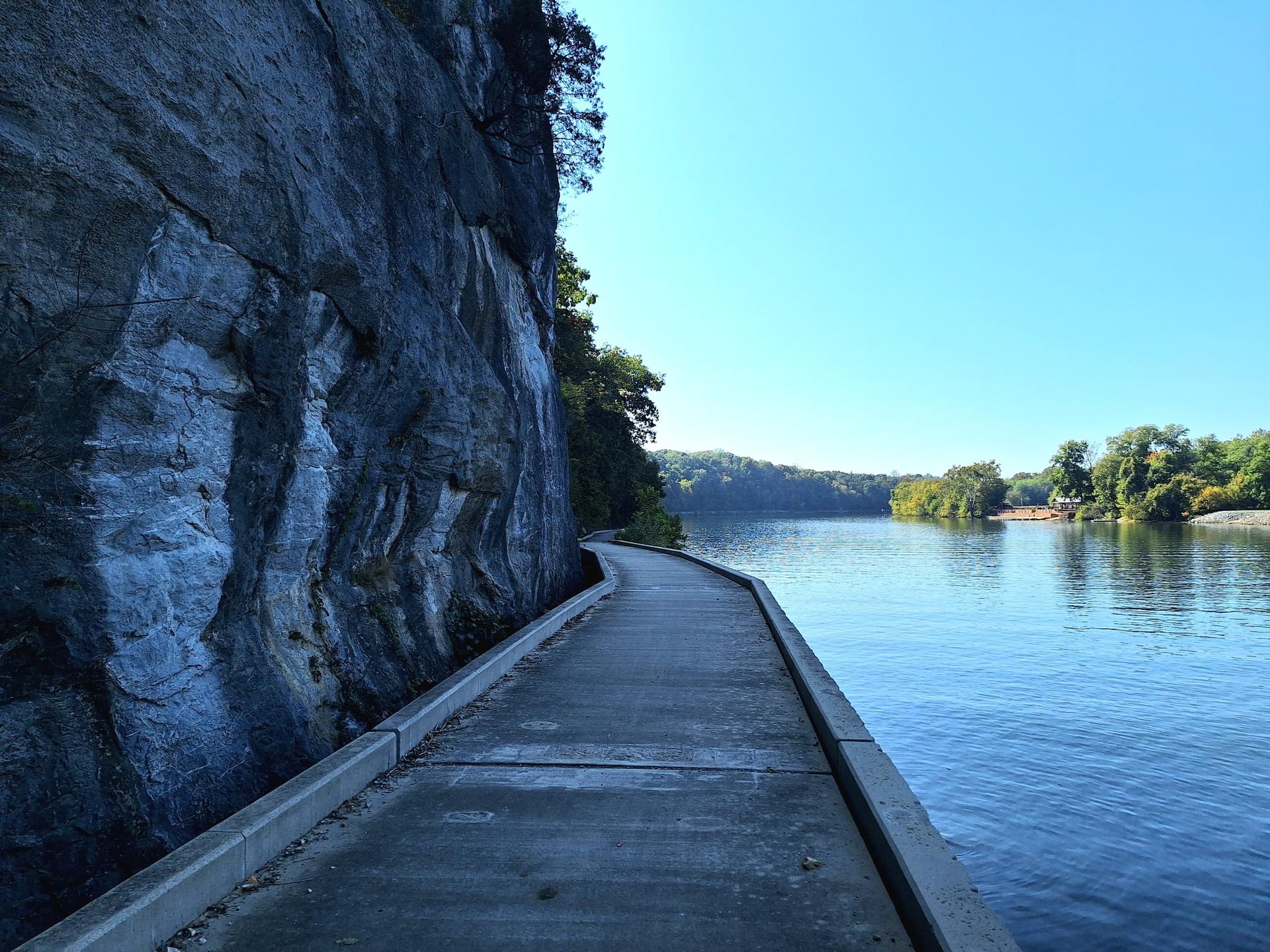 Concrete path next to a towering rock cliff and a calm river.