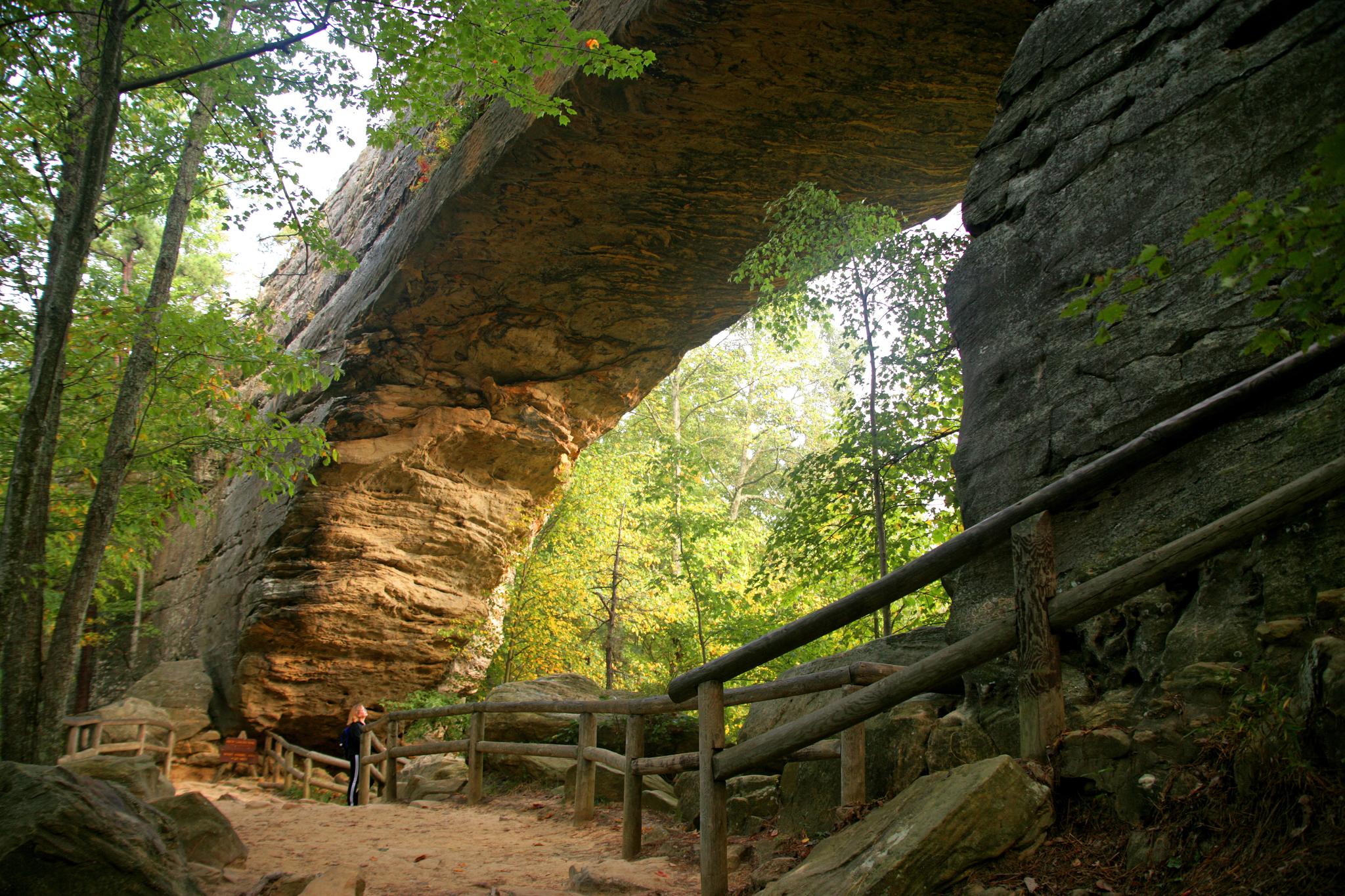 Large natural rock arch over a dirt path with wooden railings and people.