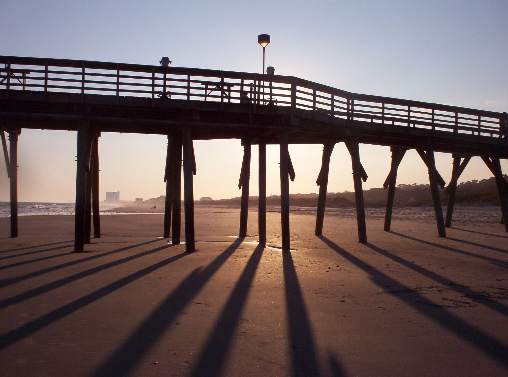 Silhouetted pier at sunset, casting long shadows across a sandy beach. - Myrtle Beach State Park & South Ocean Boulevard Beach Loop