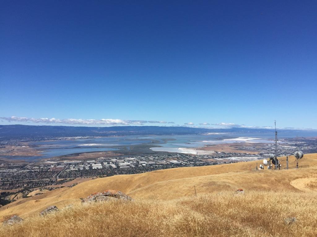 Golden hills with communication towers overlooking a city and water under a clear sky.