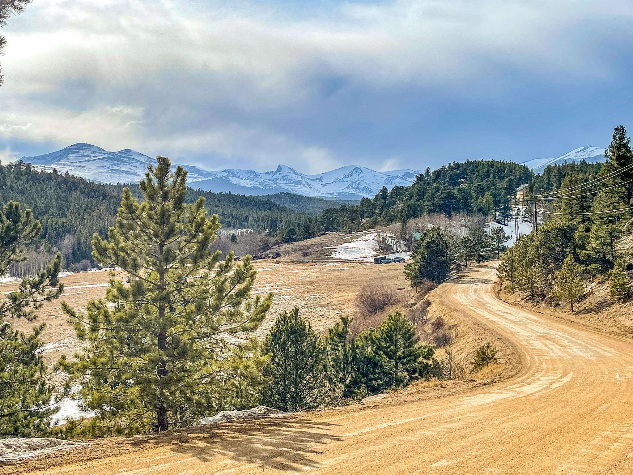 A dirt road winds through a valley with pine trees and snowy mountains. - Ceran St. Vrain Trail