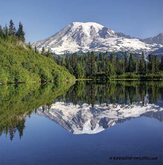 Snow-capped mountain reflecting perfectly in a calm lake, surrounded by green forest.