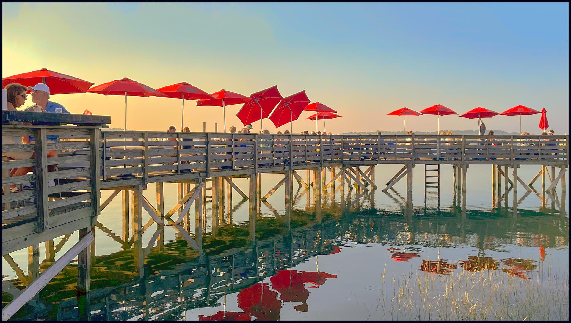 People on a long wooden pier with many red umbrellas at sunset.