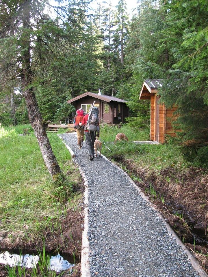 Two hikers and a dog walk on a gravel path towards a cabin in a dense forest.