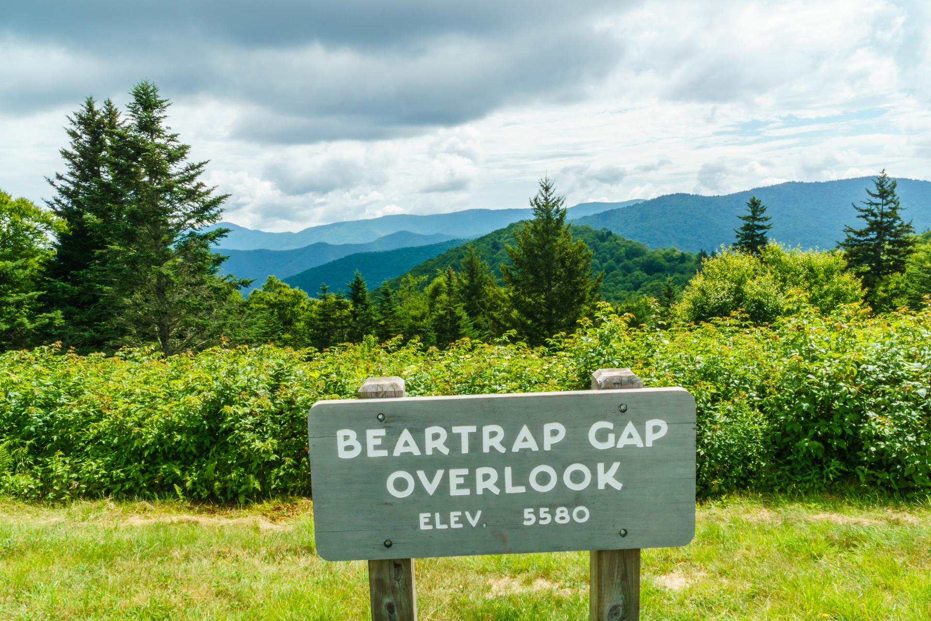 Wooden sign in foreground with green mountain landscape under cloudy sky.