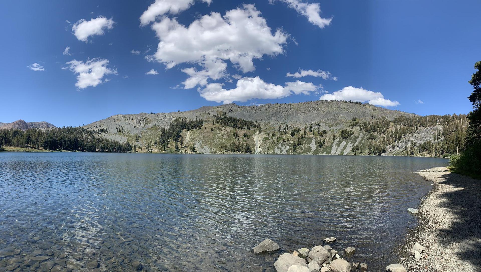Wide clear lake with a rocky shore and forested mountains under a blue sky.