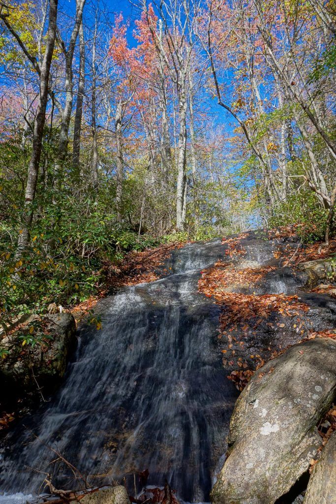 A small waterfall flows over rocks amidst vibrant autumn trees under a blue sky. - Graybeard Trail