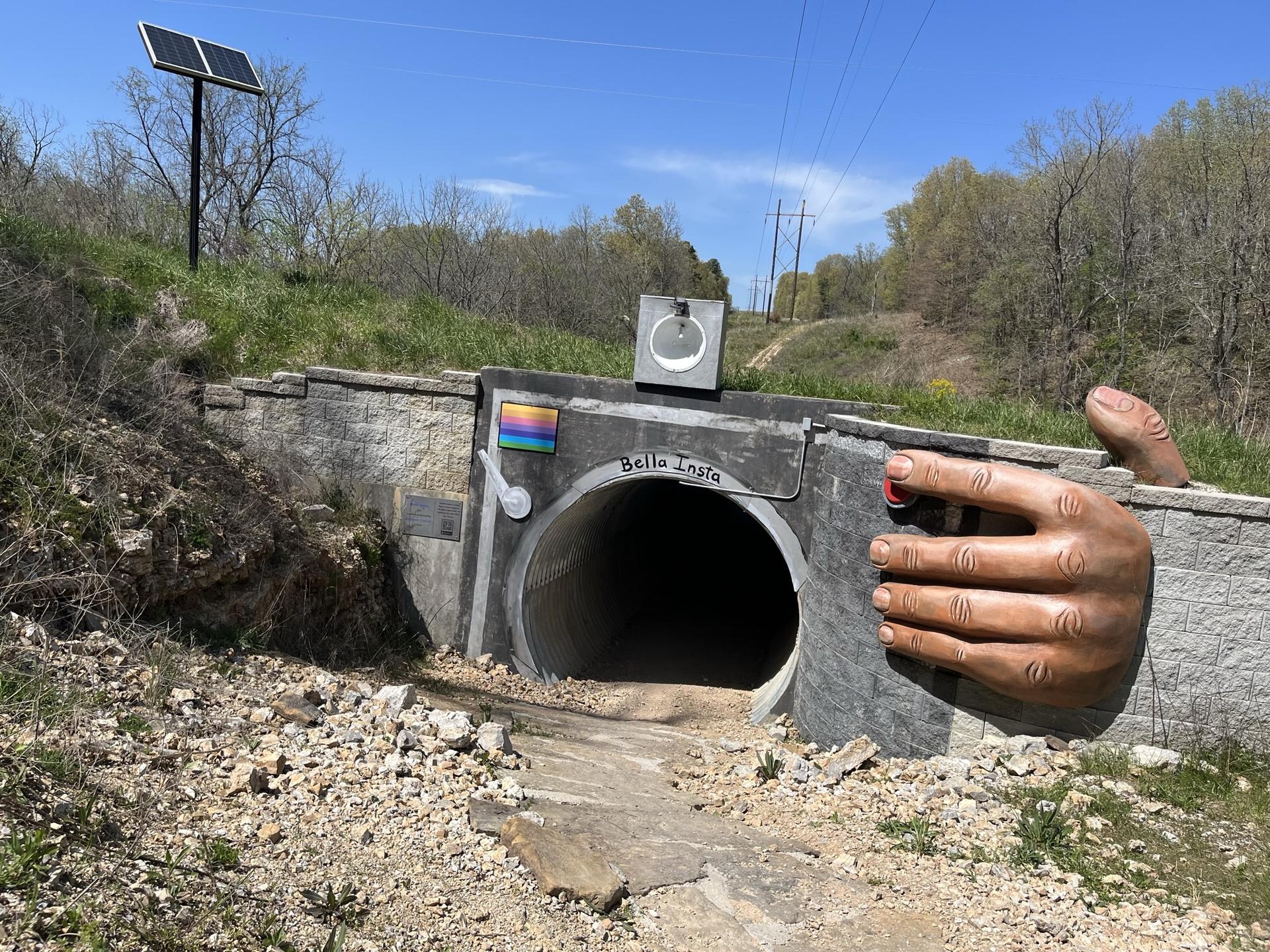 A culvert painted like a camera with a large hand, under a sunny sky.