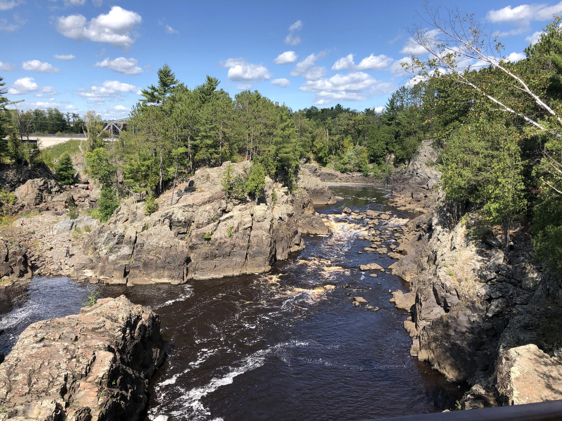 Dark river winding through a rocky gorge with green trees under a sunny sky.