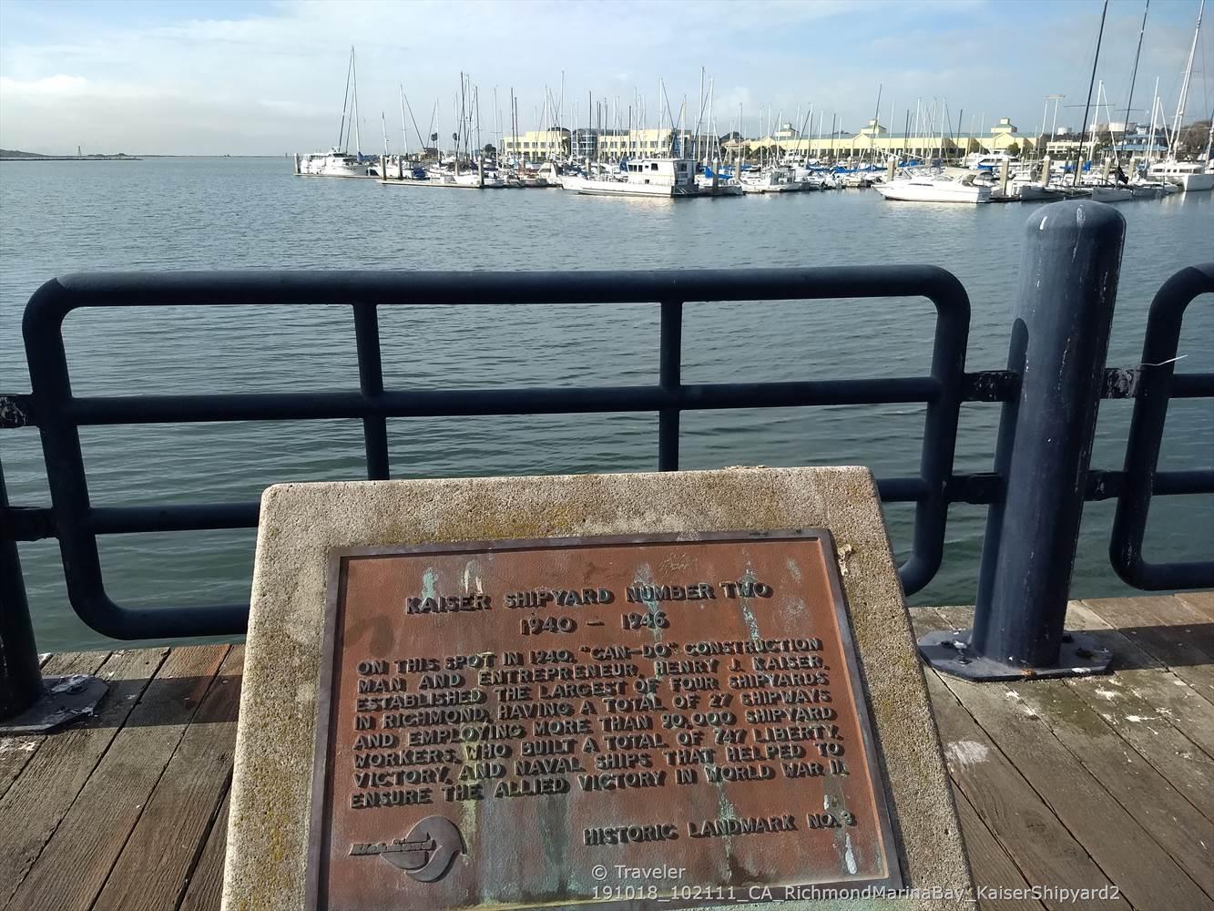 A bronze plaque on a pier railing overlooking a marina with boats.