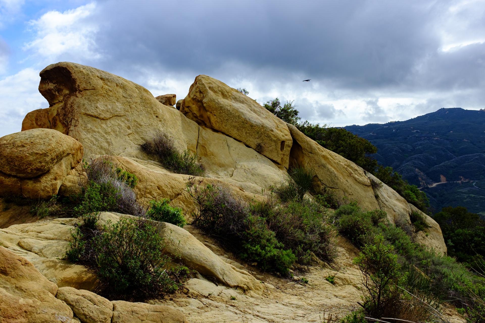 Large rock formations and scrub brush under a cloudy sky with distant mountains.