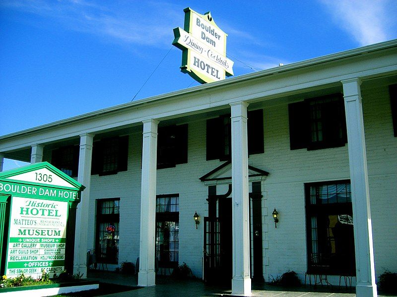 White building with columns, a large hotel sign, and a green museum sign. - Boulder Theatre – Boulder Dam Hotel loop from Clark County Museum