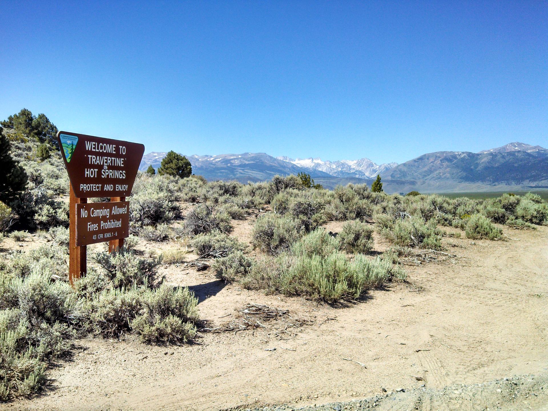 A wooden sign stands in a sunny, arid landscape with distant snow-capped mountains.