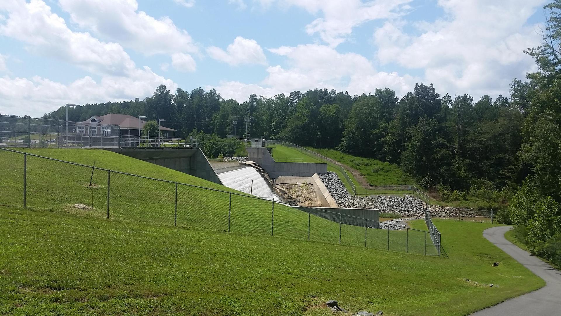 A concrete dam structure with grassy slopes and distant trees under a cloudy sky.