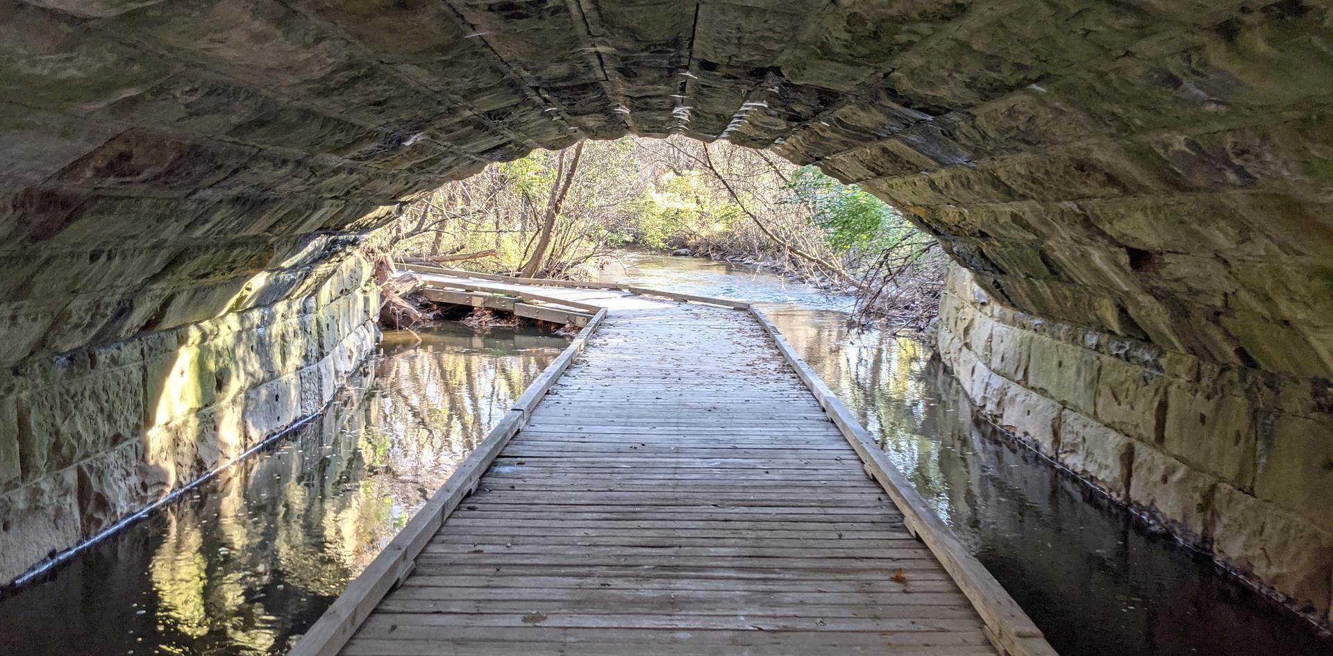 A wooden boardwalk extends through a dark stone tunnel over water.