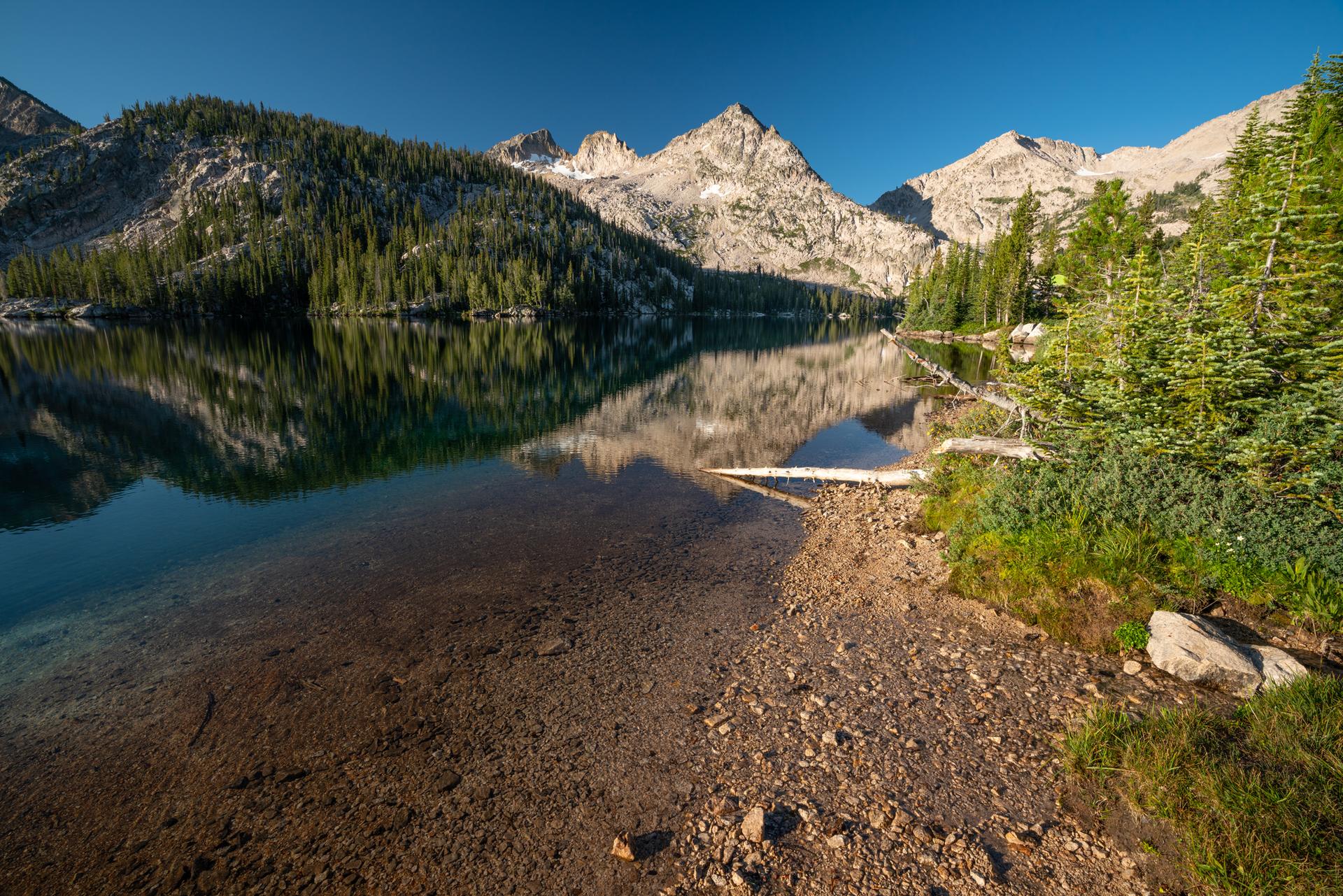 Calm lake reflecting rugged mountains and pine trees, with a rocky shore.
