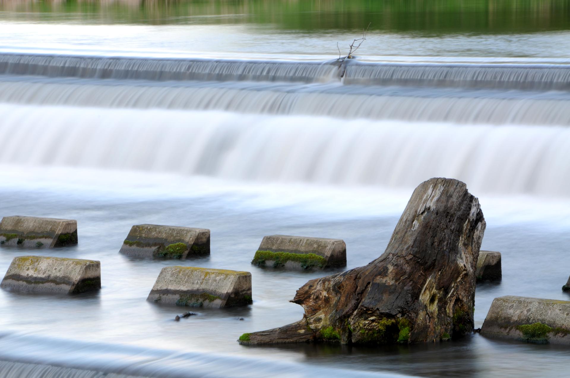 Long exposure of a waterfall with a large tree stump and concrete blocks.
