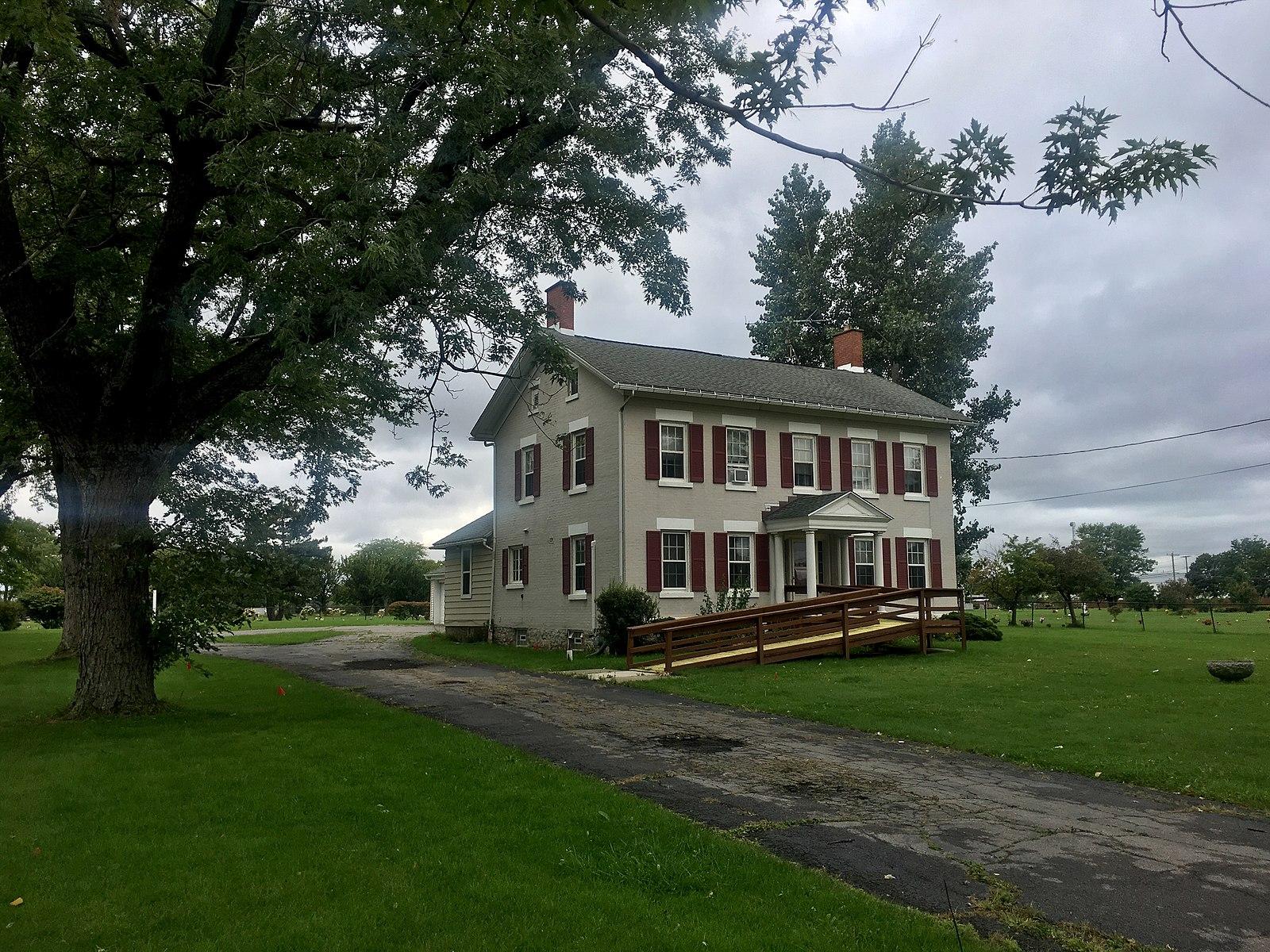 Historic house with red shutters and a ramp, surrounded by green lawn and trees.