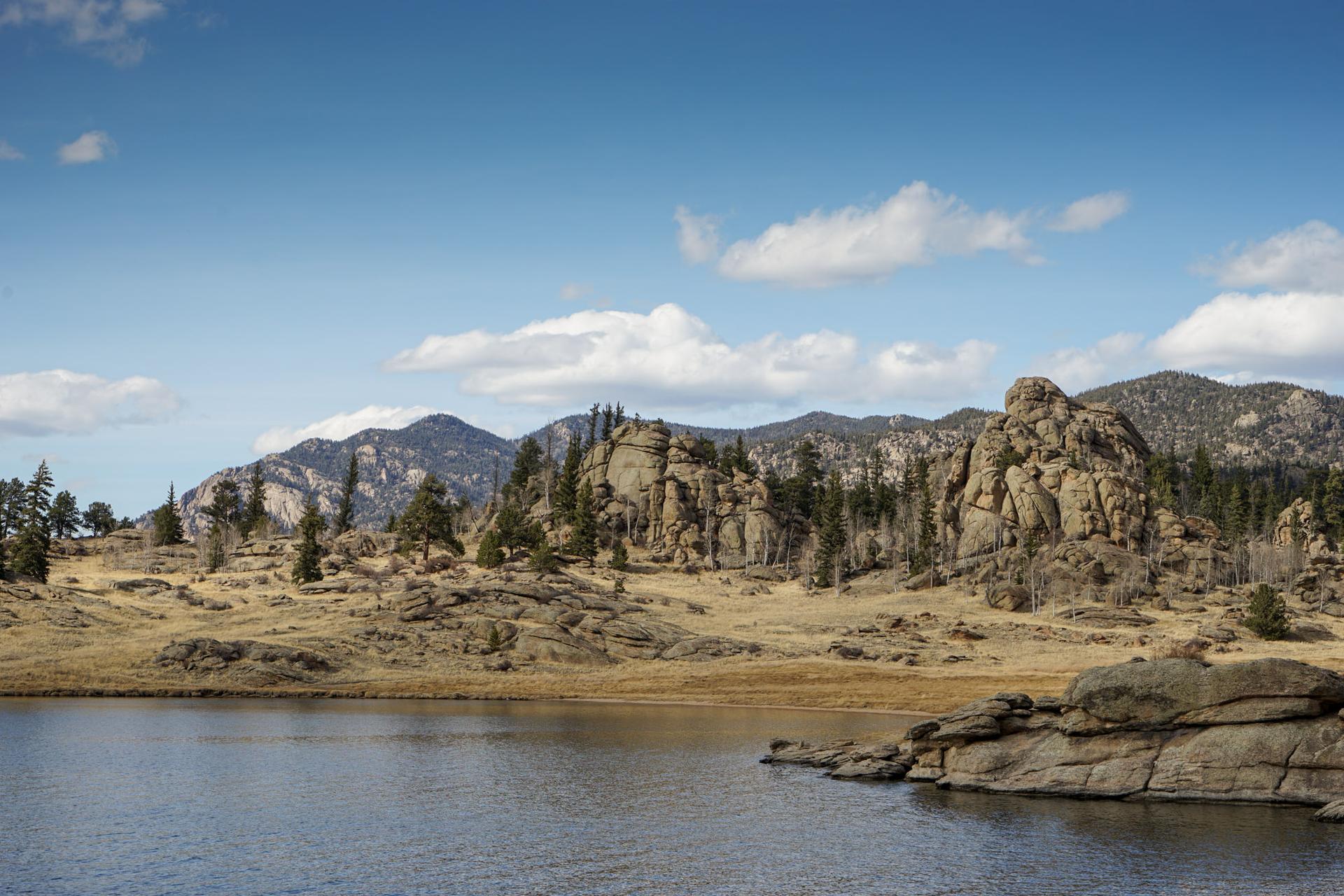 A calm lake with rocky shores, dry hills, pine trees, and distant mountains.